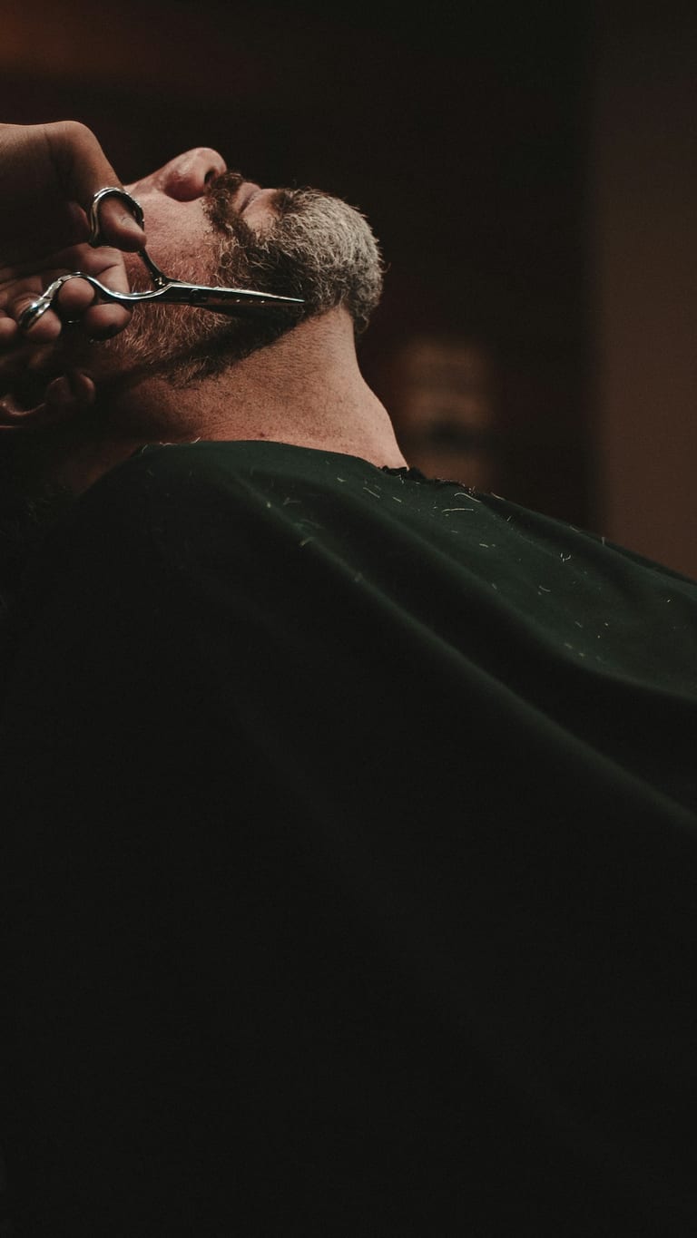 A barber trimming a reclined client’s beard with scissors